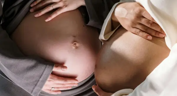 Två gravida magar, en med grå tröja, den andra med ljus. Händer vilar mjukt på magarna, en ring syns. Känslan av ligament gravid.